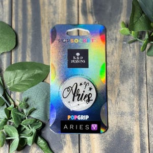 Op de afbeelding: Een ronde telefoonhouder met het woord Aries in zwarte schrijfletters, het sterrenbeeldteken Ram en sterren op een witte achtergrond. Het item is bevestigd op een rechthoekige holografische kaart met de tekst PopSockets, K&D Designs en PopGrip.