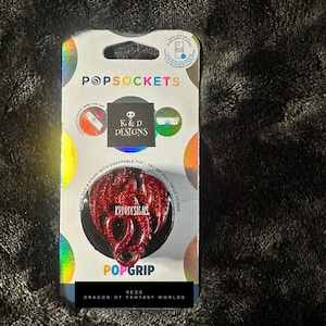 May include: A red glittery dragon-shaped phone grip and stand in its original packaging. The packaging features the brand name Popsockets, the logo for K&D Designs, and the text Reds Dragon of Fantasy Worlds. The product is a swappable Popgrip.