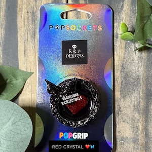 May include: A black PopSocket PopGrip with a red crystal heart design. The PopSocket is on a holographic background with the text "POPSOCKETS" and "K&D DESIGNS". The PopGrip is labeled "RED CRYSTAL".