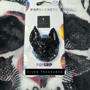 Puede incluir: PopSockets PopGrip negro con diseño de murciélago, detalles de purpurina y el texto "RIVER TREASURES". El accesorio para teléfono está sobre un respaldo blanco y colorido de la marca PopSockets.