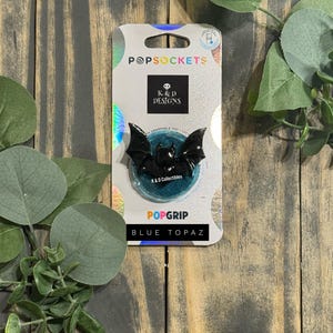 Op de afbeelding: Een zwarte, vleermuisvormige PopSocket-telefoonhouder op een witte displaykaart met holografische accenten. De kaart bevat de tekst PopSockets, K & D Designs, PopGrip en Blue Topaz. De vleermuisfiguur is glanzend zwart met puntige oren en vleugels.