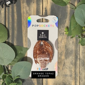 May include: A PopSockets PopGrip featuring a skull with orange glitter dragon wings on a K&D Designs backing card. The text on the card reads PopSockets, K&D Designs, K&D Collectibles, PopGrip, and Orange Topaz Dragon. The accessory is orange.