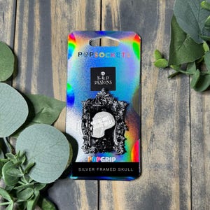 May include: A silver framed skull PopSocket on a holographic background. The PopSocket features a detailed skull design within an ornate frame. The packaging includes the text "K&D Designs Collectibles" and "Silver Framed Skull."