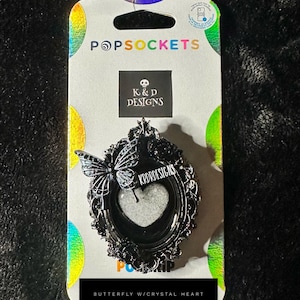 May include: A black PopSockets phone grip with a butterfly and crystal heart design. The grip is in a decorative frame with the text "K&D Designs" and "Butterfly w/Crystal Heart". The packaging has the PopSockets logo.
