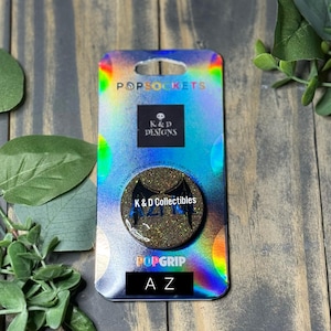 Op de afbeelding: Een ronde telefoonhouder met een donker, glinsterend oppervlak, zwarte vleugelafbeeldingen en de tekst K & D Collectibles en A Z, bevestigd op een rechthoekige, holografische achterkaart met het opschrift PopSockets en K & D Designs.