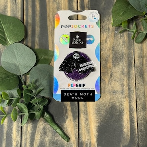 Op de afbeelding: Een PopSockets PopGrip telefoonaccessoire met een zwart doodshoofdvlinder-embleem met schedeldetail, gecentreerd op een ronde paarse glitterbasis. Het product is bevestigd op de originele verpakking, met de tekst K&D DESIGNS en DEATH MOTH MUSE.