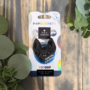 Op de afbeelding: Een zwart, vleermuisvormig PopGrip-telefoonaccessoire op de originele verpakking. De verpakking bevat de tekst PopSockets, K&D Designs en Glow-in-the-Dark Sky Blue. De vleermuisfiguur heeft een glanzende afwerking en puntige oren.