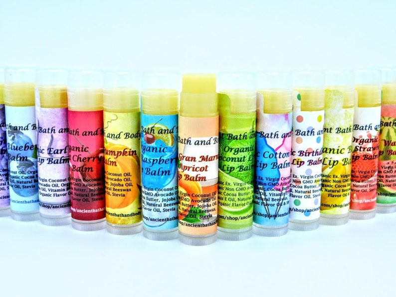 May include: A row of 12 colorful lip balms in tubes with various fruit and floral scents. The lip balms are labeled with the brand name "Bath and Body" and the scent name. The lip balms are arranged in a rainbow pattern.