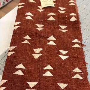 May include: A rectangular piece of woven fabric in a reddish-brown hue, adorned with repeating white triangle patterns. The fabric has a fringed edge and a textured appearance, suggesting a rustic or handmade quality. A small sticky note is attached.