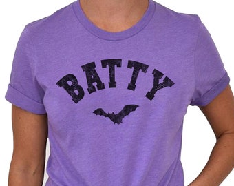 Puede incluir: Una camiseta morada con la palabra "BATTY" en negro, fuente desgastada, con un gráfico de murciélago negro debajo.
