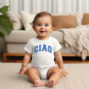 May include: A baby wearing a white onesie with the word "CIAO" printed in blue on the front. The baby is sitting on a white rug in front of a couch.