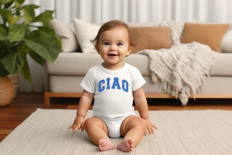 May include: A baby wearing a white onesie with the word "CIAO" printed in blue on the front. The baby is sitting on a white rug in front of a couch.