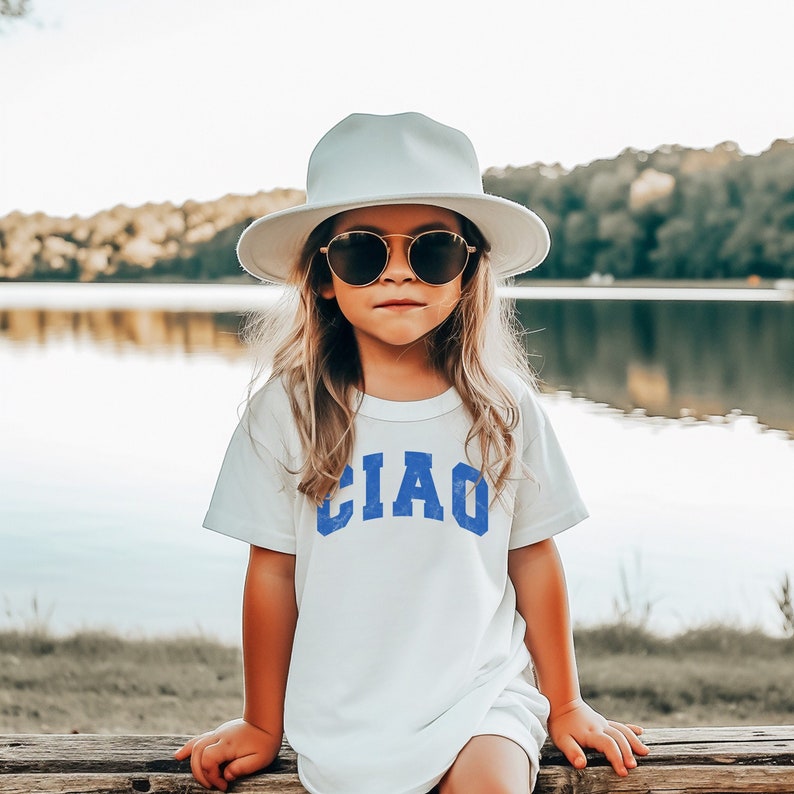 May include: A young person wearing a white t-shirt with the word "CIAO" printed in blue. They are wearing a white sun hat and sunglasses.