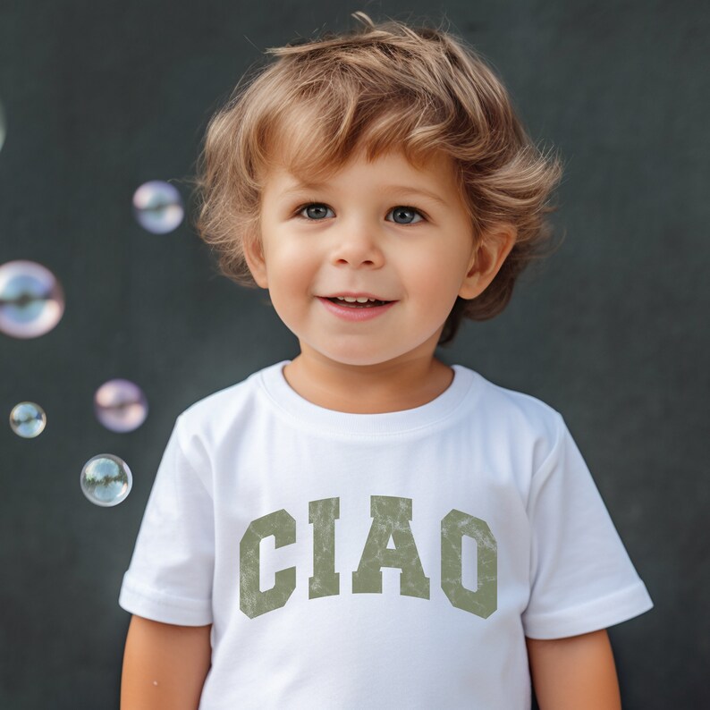 May include: A young child wearing a white t-shirt with the word "CIAO" printed in green on the front. The child is smiling and looking to the right. There are soap bubbles in the background.