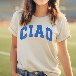 May include: A white t-shirt with the word "CIAO" printed in blue letters. The letters are in a vintage, distressed style.