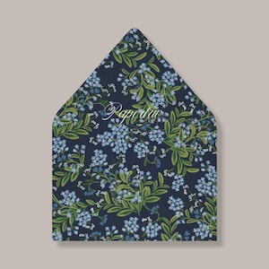 May include: An envelope with a floral pattern featuring blue flowers and green leaves on a dark navy background. The word "Paperie" is written in a cursive font across the front of the envelope.