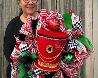 May include: A festive Christmas wreath featuring a red, black, and white color scheme. The wreath includes a red top hat with gold trim, decorative ribbons, and ornaments. The text "Four Thirteen Design LLC" is visible on the hat.