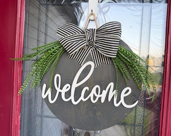 May include: A circular dark wood door sign featuring the white script text "welcome" in the center. The sign is decorated with a black and white striped fabric bow at the top and two sprigs of green artificial fern leaves extending outward.