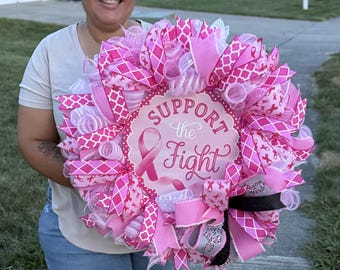 May include: A circular decorative wreath featuring pink and white mesh ribbons, patterned ribbons with breast cancer awareness symbols, and a central sign with a pink ribbon graphic and the text Support the Fight in pink script lettering.