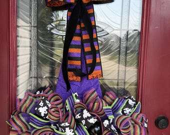 May include: A Halloween-themed witch hat wreath featuring a purple fabric base adorned with layers of mesh ribbon in black, purple, orange, and lime green. The wreath is topped with a large bow made of striped orange and purple glittered ribbon.