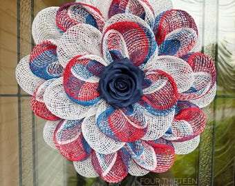 May include: A circular wreath constructed from red, white, and blue mesh loops arranged in a flower-like pattern. A single dark navy blue artificial rose is positioned in the center. The text FOUR THIRTEEN DESIGN is visible at the bottom right.