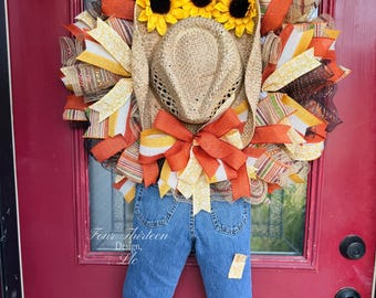 May include: A scarecrow-themed wreath featuring a straw cowboy hat, three yellow sunflowers, and blue denim jeans. The wreath is decorated with orange, yellow, and brown mesh ribbons and includes a patch on the jeans. Text reads Four Thirteen Design, Llc.
