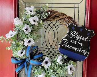May include: A decorative wreath featuring a brown twig base, white anemone flowers with dark centers, and green foliage. A blue and black striped ribbon is attached, along with a black shield-shaped sign reading Blessed ARE THE Peacemakers.