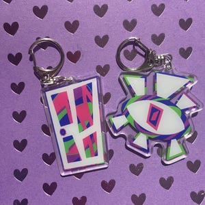 Puede incluir: Dos llaveros acrílicos con llaveros plateados. Un llavero es un rectángulo con un diseño de puerta en rosa, morado, verde y azul. El otro es un diseño blanco, verde y morado con forma de ojo central. El fondo es morado con forma de corazón.