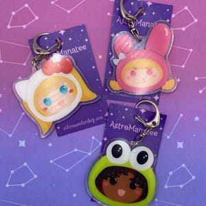 May include: Three acrylic keychains featuring cartoon characters in animal-themed hoods. One wears a white cat hood with a red bow, one a pink rabbit hood, and one a green frog hood. Each is attached to a silver metal ring on a purple backing card.