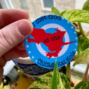 May include: A blue and red circular sticker with white stars around the edge. The sticker says "I met God at the Chicago Rat Hole".