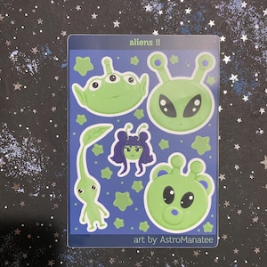 May include: A sticker sheet featuring various green alien designs on a blue background. The sheet includes aliens with three eyes, antennae, and bear-like features, along with green star shapes. Text reads "aliens !!" and "art by AstroManatee."