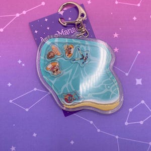 May include: A clear acrylic shaker keychain featuring a pool scene with a light blue water surface, a yellow sandy edge, and four small floating character charms. The keychain includes a silver metal lobster clasp and a purple backing card.
