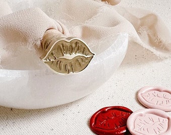 May include: A gold-toned lip-shaped wax seal stamp with five red and pink wax seals. The wax seals are imprinted with a lip design.
