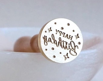 May include: A gold-toned metal stamp with the words "Happy Birthday" engraved on it. The stamp is surrounded by small dots and stars.