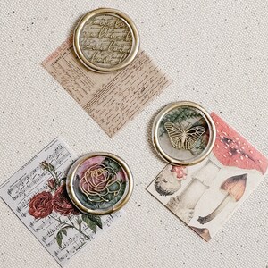 May include: Three decorative wax seals with gold accents. The first seal features a floral design with a rose and green leaves. The second seal features a butterfly with gold wings. The third seal features a handwritten message in gold lettering.