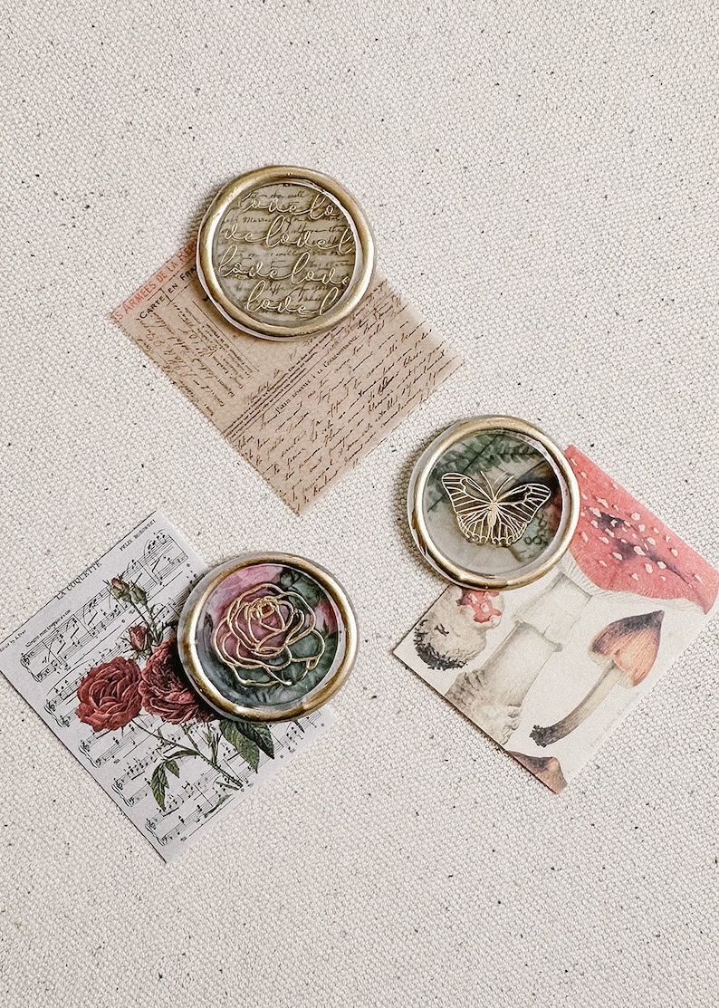 May include: Three decorative wax seals with gold accents. The first seal features a floral design with a rose and green leaves. The second seal features a butterfly with gold wings. The third seal features a handwritten message in gold lettering.