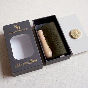 May include: A black box with a gold logo that says "Made of Honour" and "Wax seal stamp". The box is open and shows a wooden handle wax seal stamp, a green velvet pouch, and a gold wax seal with a leaf design.