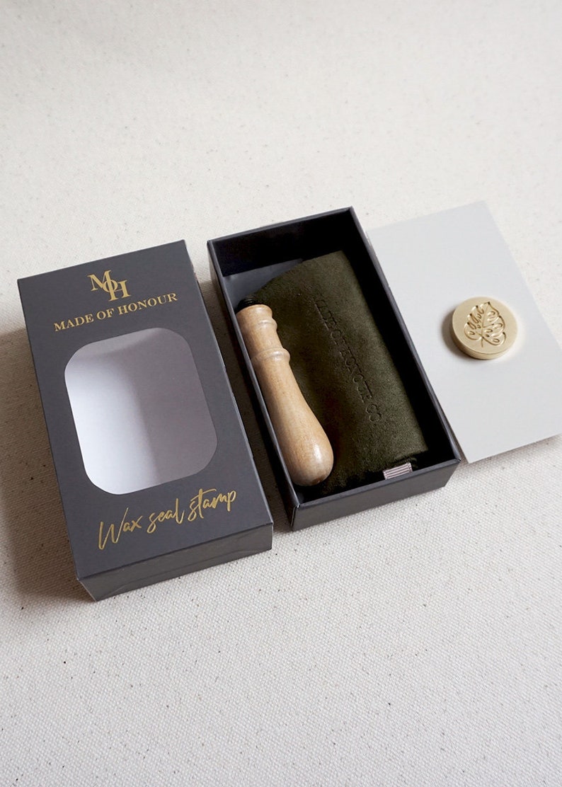 May include: A black box with a gold logo that says "Made of Honour" and "Wax seal stamp". The box is open and shows a wooden handle wax seal stamp, a green velvet pouch, and a gold wax seal with a leaf design.