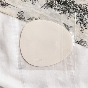 May include: A white, oval-shaped, blank card with the text "HONOUR LONDON" printed in black on the bottom. The card is sitting on a white, textured surface.
