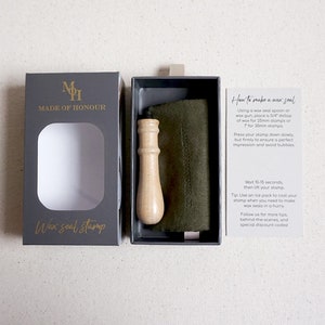May include: A black box with a gold logo that says "Made of Honour" and the words "Wax seal stamp" in gold lettering. The box is open and shows a wooden handle wax seal stamp. The stamp is in a black box with a green felt pad. A white card with instructions on how to use the wax seal stamp is next to the box.
