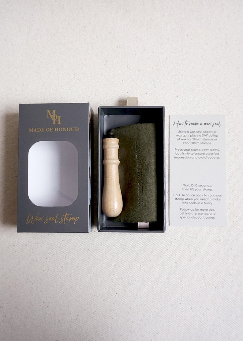 May include: A black box with a gold logo that says "Made of Honour" and the words "Wax seal stamp" in gold lettering. The box is open and shows a wooden handle wax seal stamp. The stamp is in a black box with a green felt pad. A white card with instructions on how to use the wax seal stamp is next to the box.