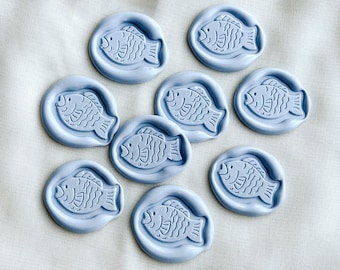 May include: Nine light blue wax seals with a fish design. The seals are arranged in a circle on a white fabric background.