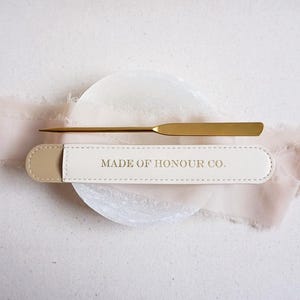 May include: A gold-colored letter opener with a matching case. The case is off-white with a beige end and the words "MADE OF HONOUR CO." printed in gold. The letter opener is lying on a white surface with a pink ribbon.