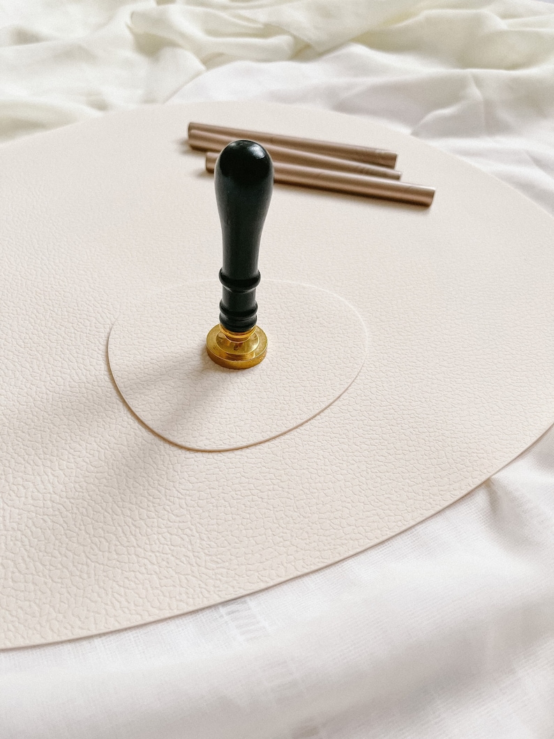 May include: A black and gold wax seal stamp with a wooden handle sits on a cream-colored surface. Three gold wax sticks are visible in the background.