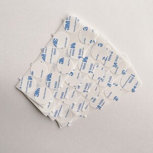 May include: A close-up of a pile of white, round, adhesive stickers with blue text that reads "3M 9448A Double Coated Tissue Tape".