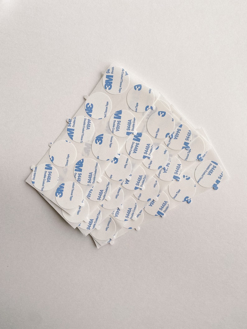 May include: A close-up of a pile of white, round, adhesive stickers with blue text that reads "3M 9448A Double Coated Tissue Tape".