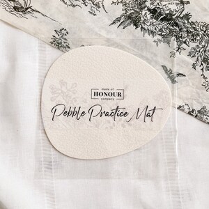 May include: A round, off-white, textured pebble practice mat with the text "Pebble Practice Mat" and "made of HONOUR company" printed in black.