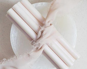 May include: A collection of four pale pink sealing wax sticks tied with a matching frayed ribbon, resting on a white, textured surface. The wax sticks are cylindrical and smooth, suggesting a craft or stationery product.