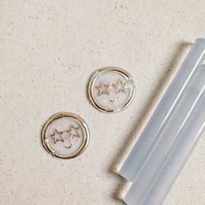 May include: Two round, white wax seals with gold trim and a design of three gold stars. The seals are on a light beige canvas background. Three clear glue sticks are in the bottom right corner of the image.