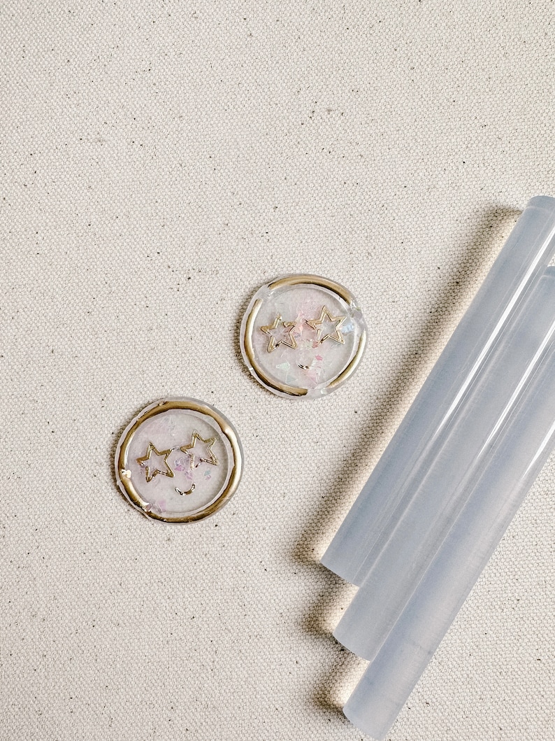 May include: Two round, white wax seals with gold trim and a design of three gold stars. The seals are on a light beige canvas background. Three clear glue sticks are in the bottom right corner of the image.